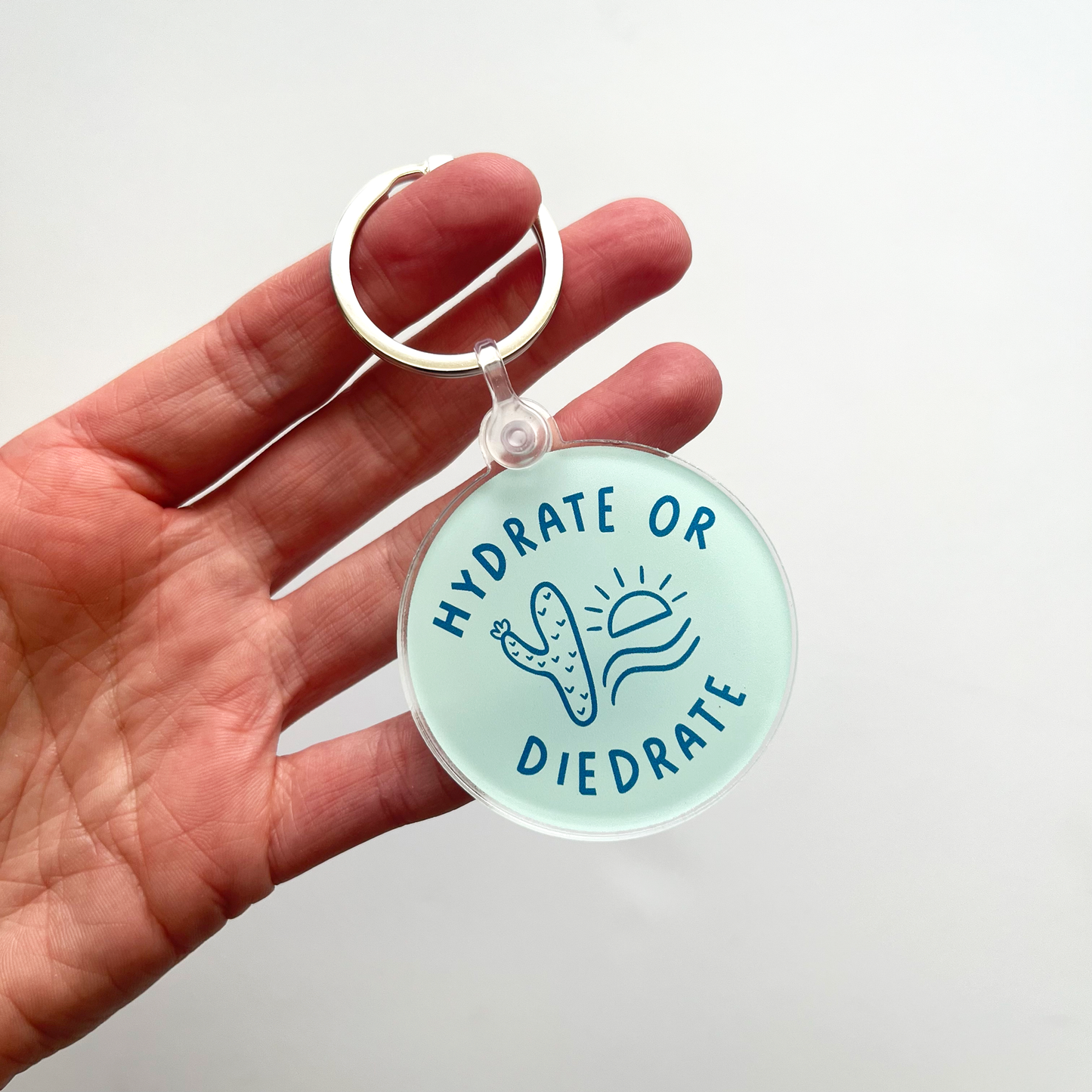 blue circle keychain with dark blue text saying hydrate or diedrate. Cactus and a sun are doodled in the middle of the circle.