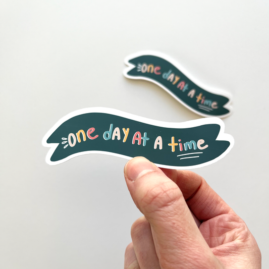 One day at a time sticker with a dark navy banner and colorful text on the inside