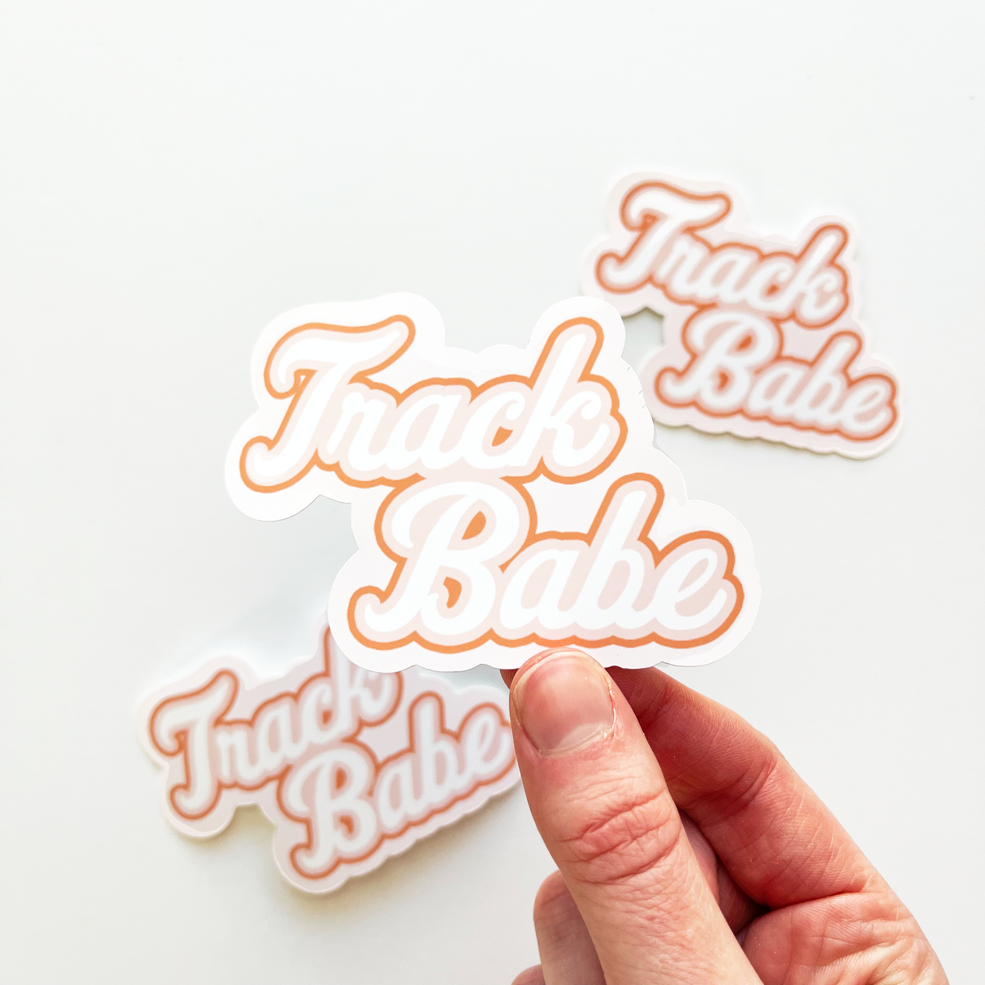 Track babe sticker with white text and outlined in peachy pink 