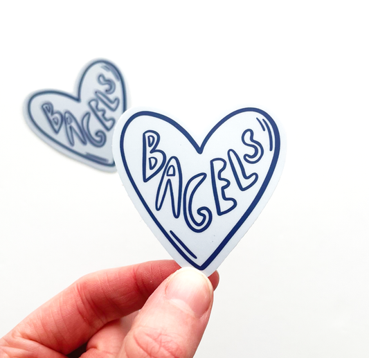 Hand holding heart shaped sticker with word bagels on the inside. The sticker is navy and light blue.