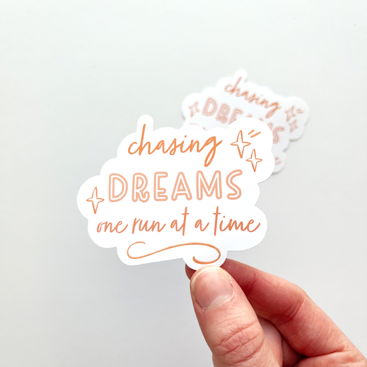 Chasing dreams one run at a time sticker with orange and pink text and a white background. Doodles of stars and lines decorate around the text