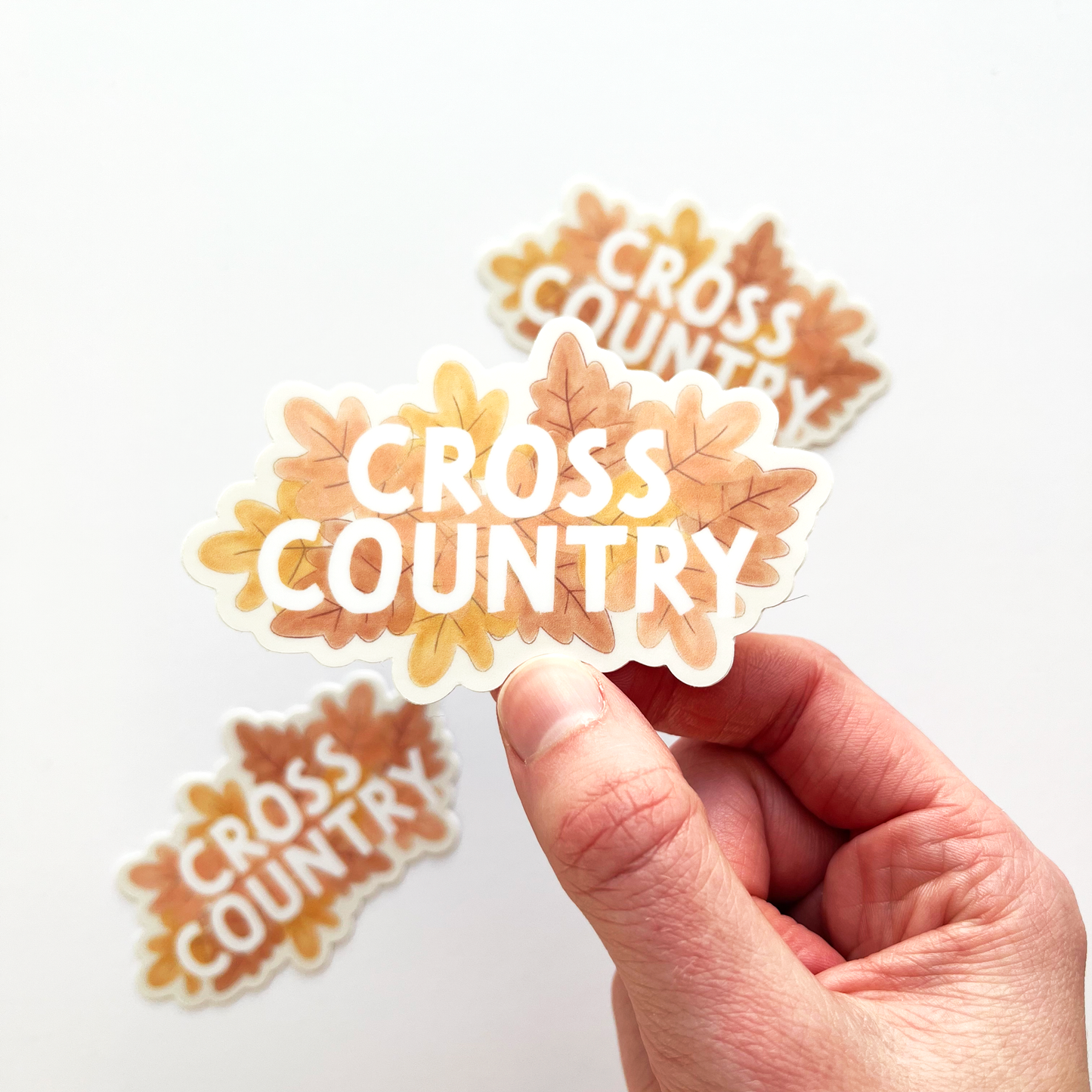 Cross Country sticker with white text and a leafy background with colors of light pink, peach and orange