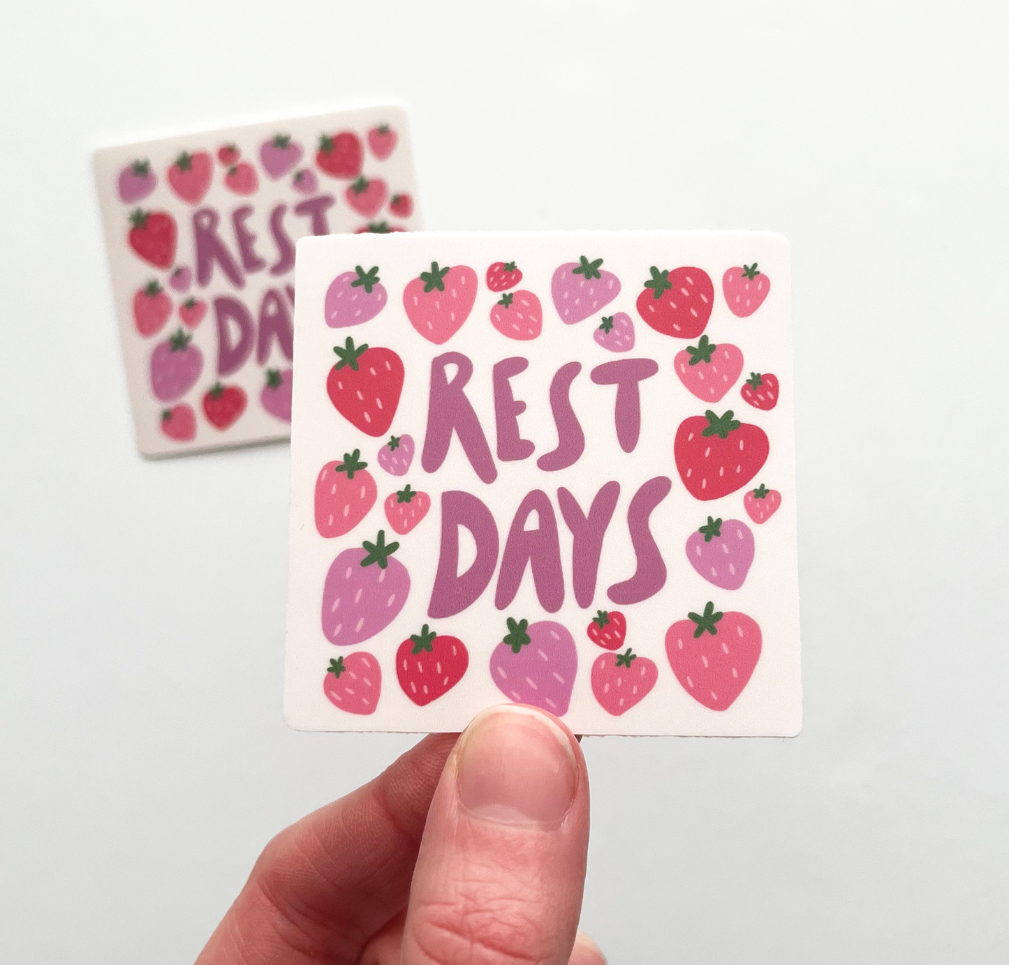 Hand holding rest days sticker. Rest Days is written in the middle of sticker in purple bubble letters. The text is surrounded by pink and purple strawberries filling the entire square.