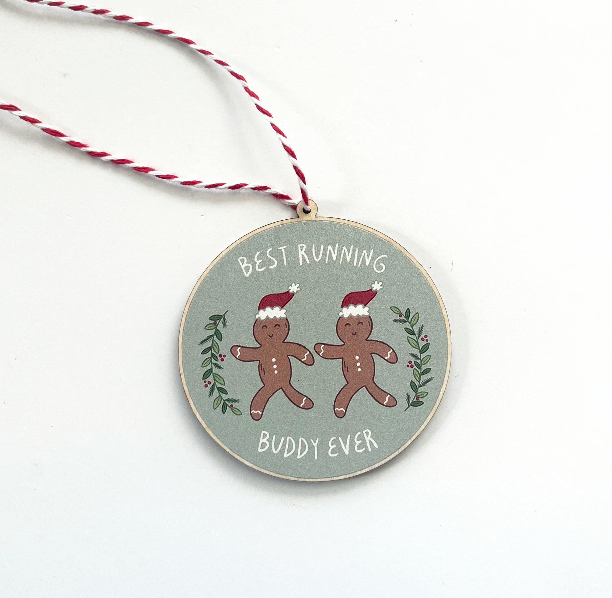 Circular ornament with two running gingerbread people with santa hats, surrounded with greenery on either side and the text best running buddy ever