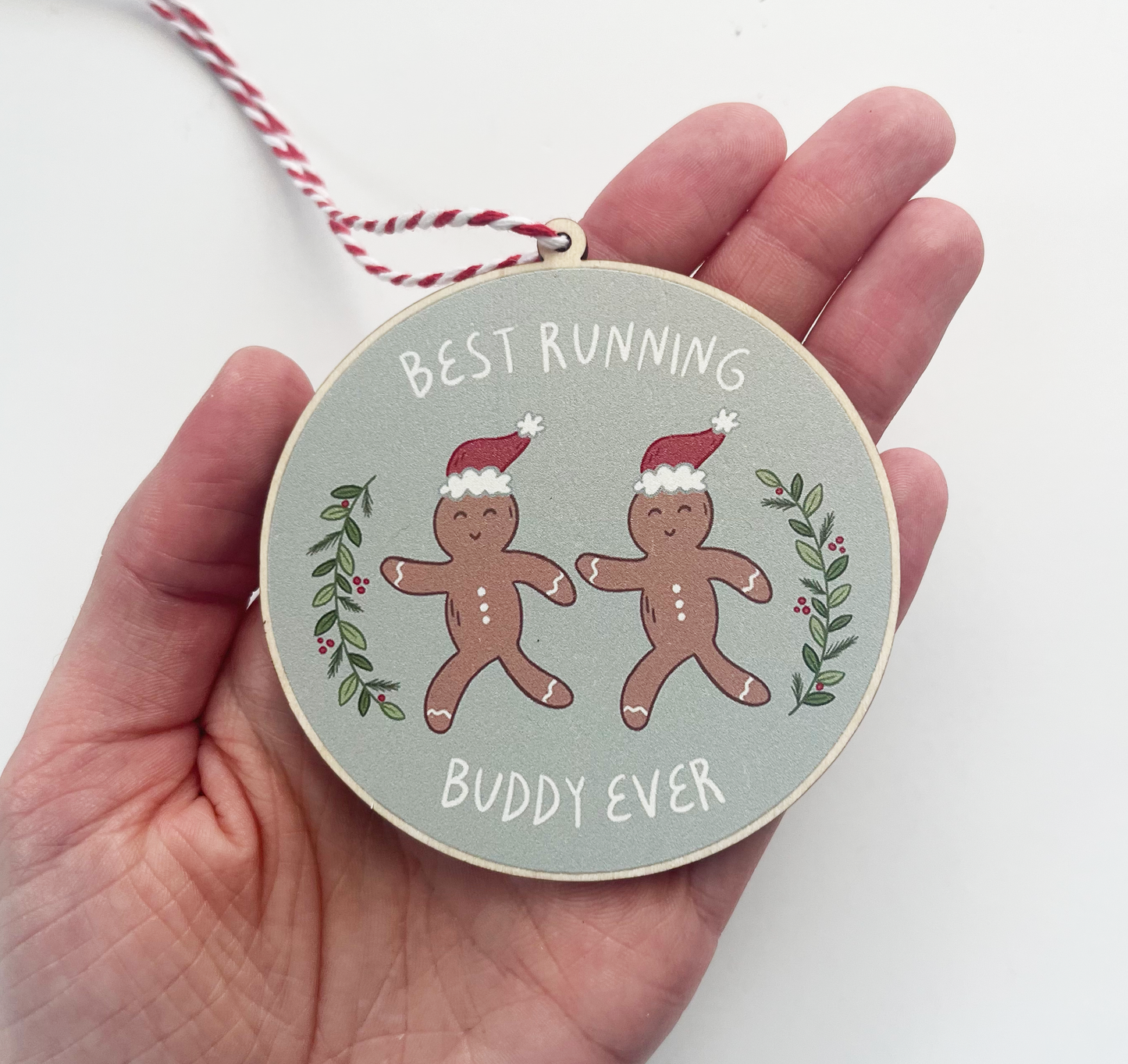 Circular ornament with two running gingerbread people with santa hats, surrounded with greenery on either side and the text best running buddy ever