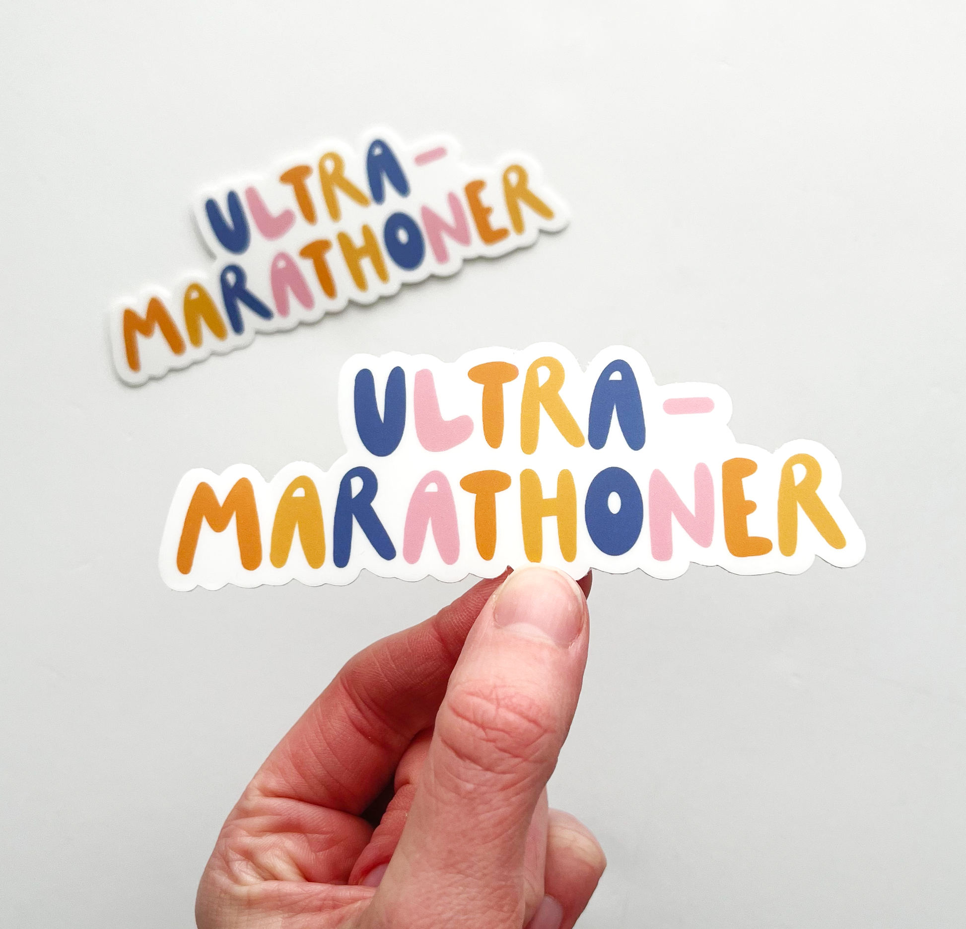 Hand holding ultramarathoner sticker. Sticker is written with bubble letters and alternating colors of navy, pink, orange and yellow.
